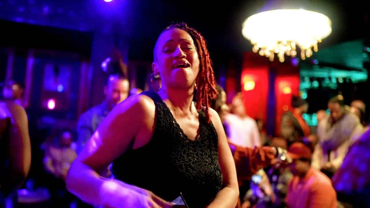 Person in black top dancing under purple lighting, chandelier and crowd in background