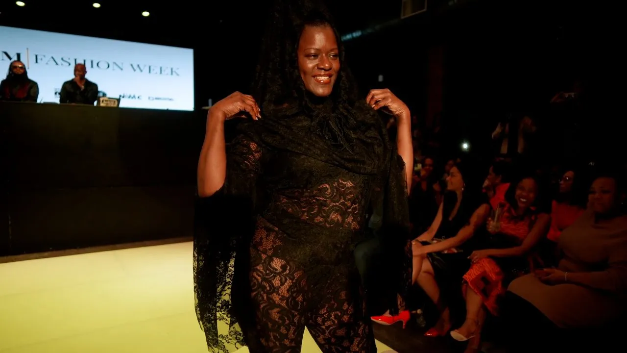 Model in black lace outfit poses on runway, Harlem Fashion Week sign, seated crowd