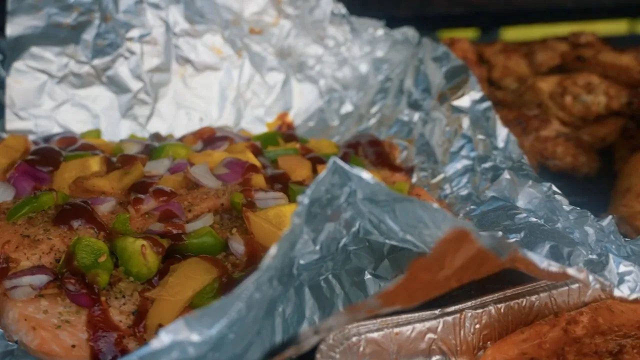 Close-up of grilled food and vegetables in foil pans, outdoor daytime lighting