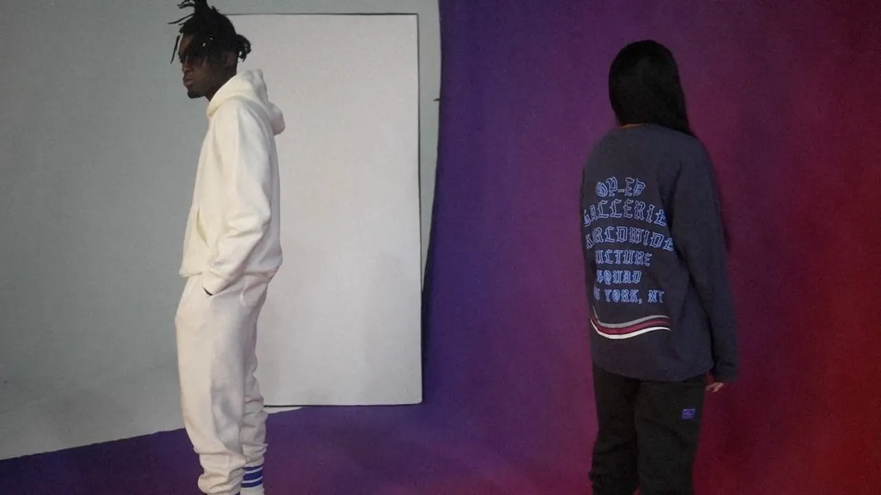 Two people in oversized hoodies against purple wall, graphic sweatshirt text visible