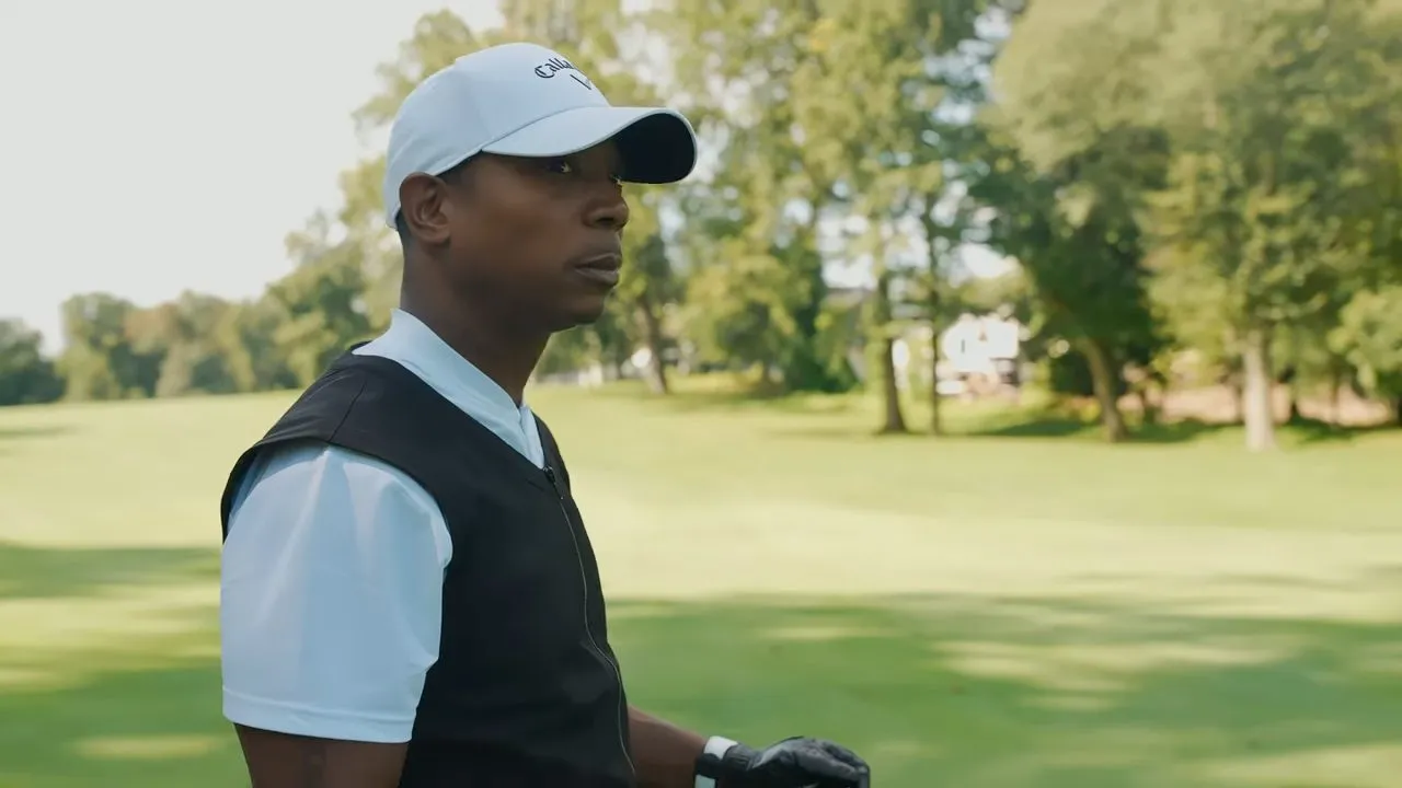 Person in white cap and vest standing on green golf course, trees behind