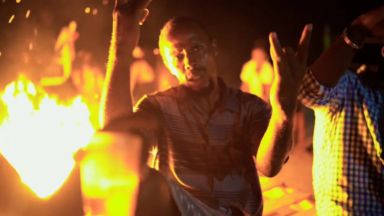 Person gesturing before bright flames at night, warm firelight, others nearby