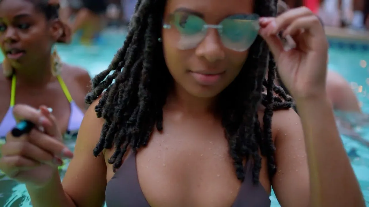 Person in sunglasses at poolside with others in swimwear, bright daylight