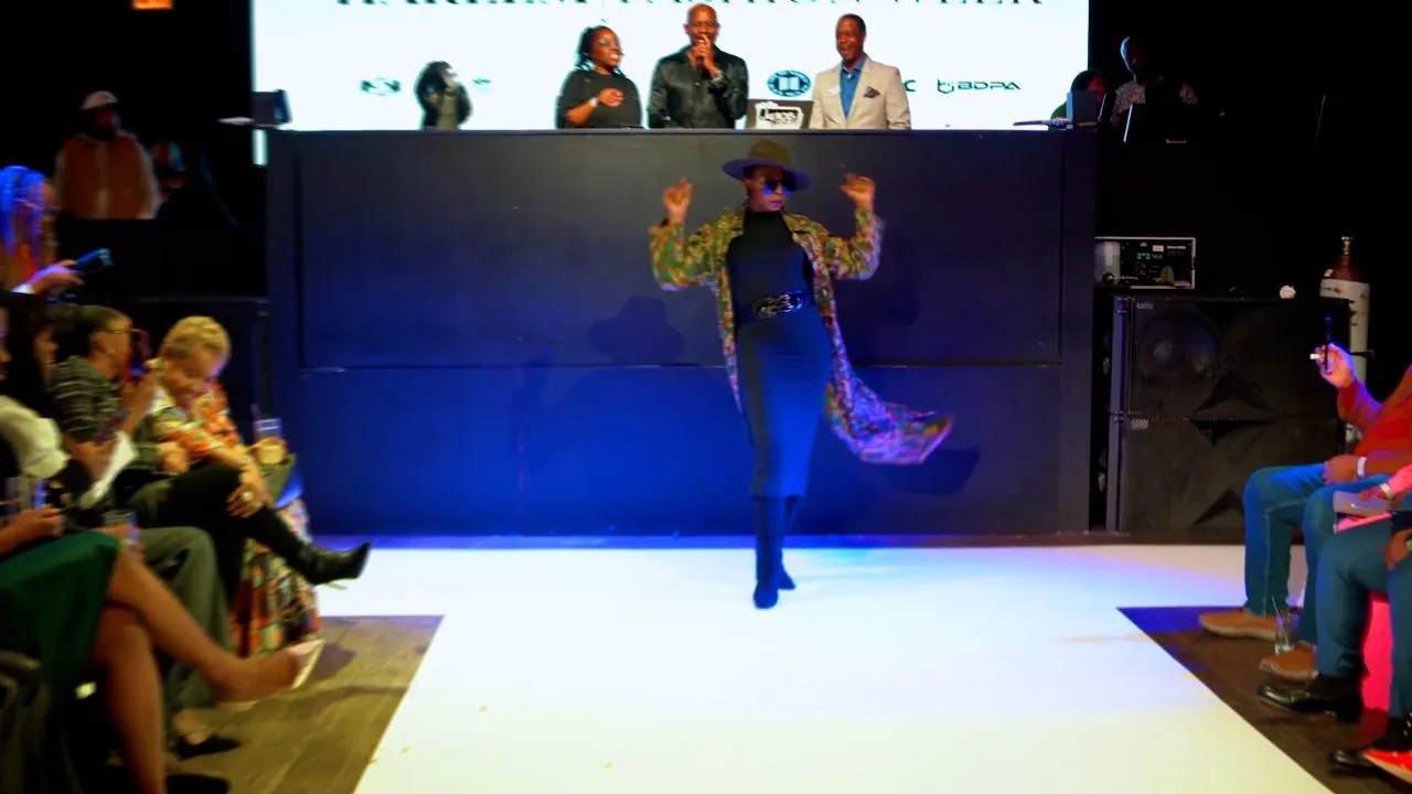 Model in patterned jacket walks white runway toward judges panel with audience alongside