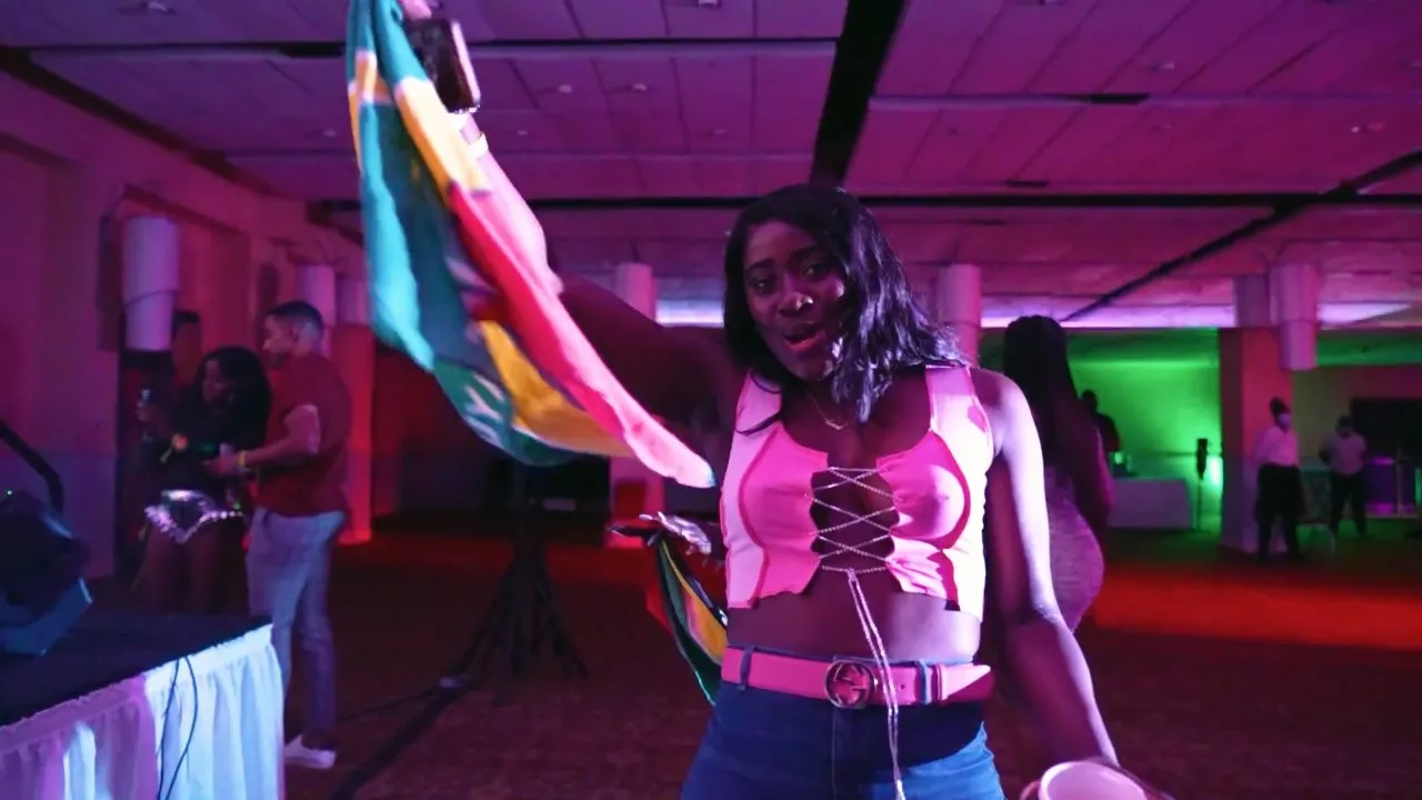 Woman in pink top holds Jamaican flag at indoor party, purple lighting