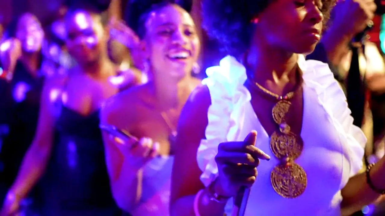 People in white celebrating with gold jewelry under colorful party lights, festive crowd