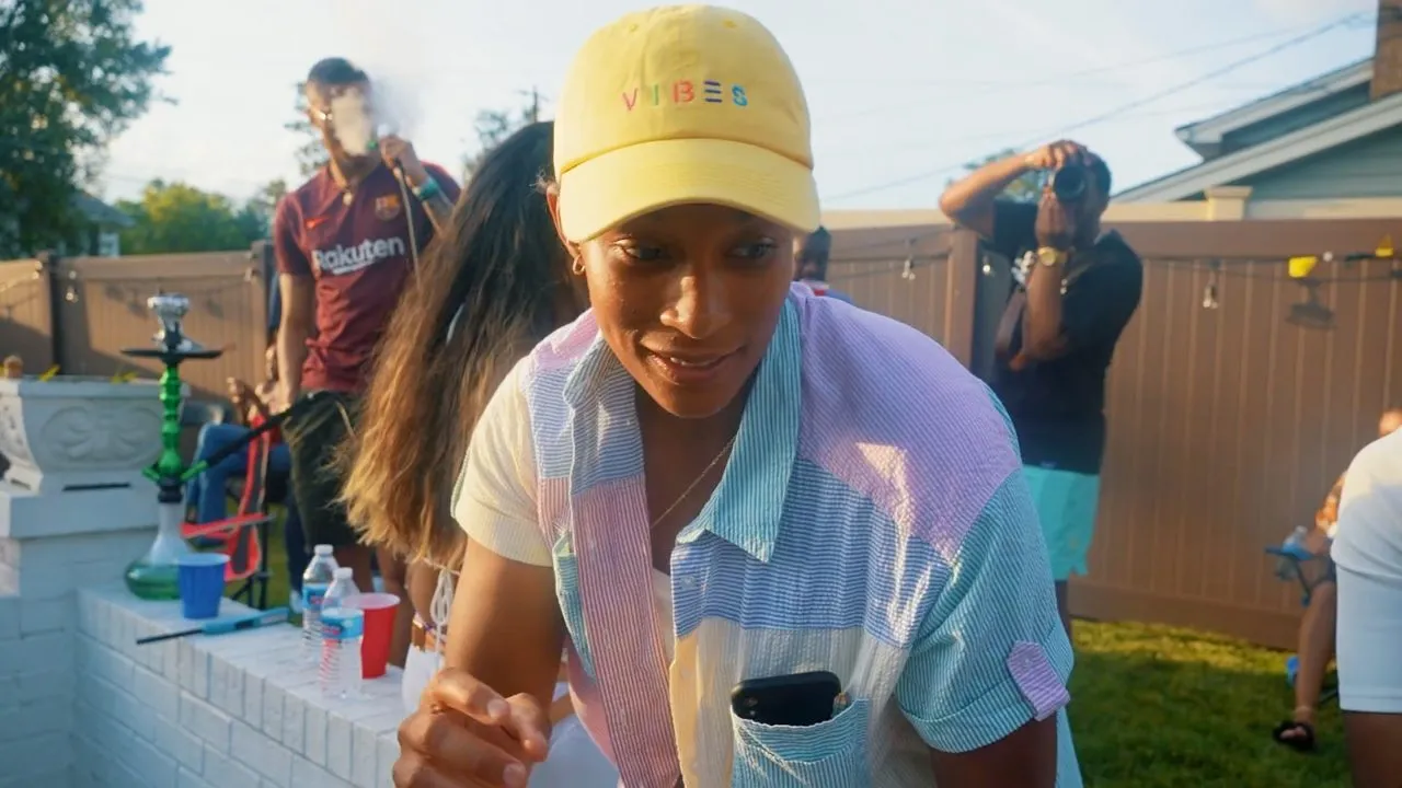 Person in yellow cap and colorful shirt dancing outdoors near fence, daytime