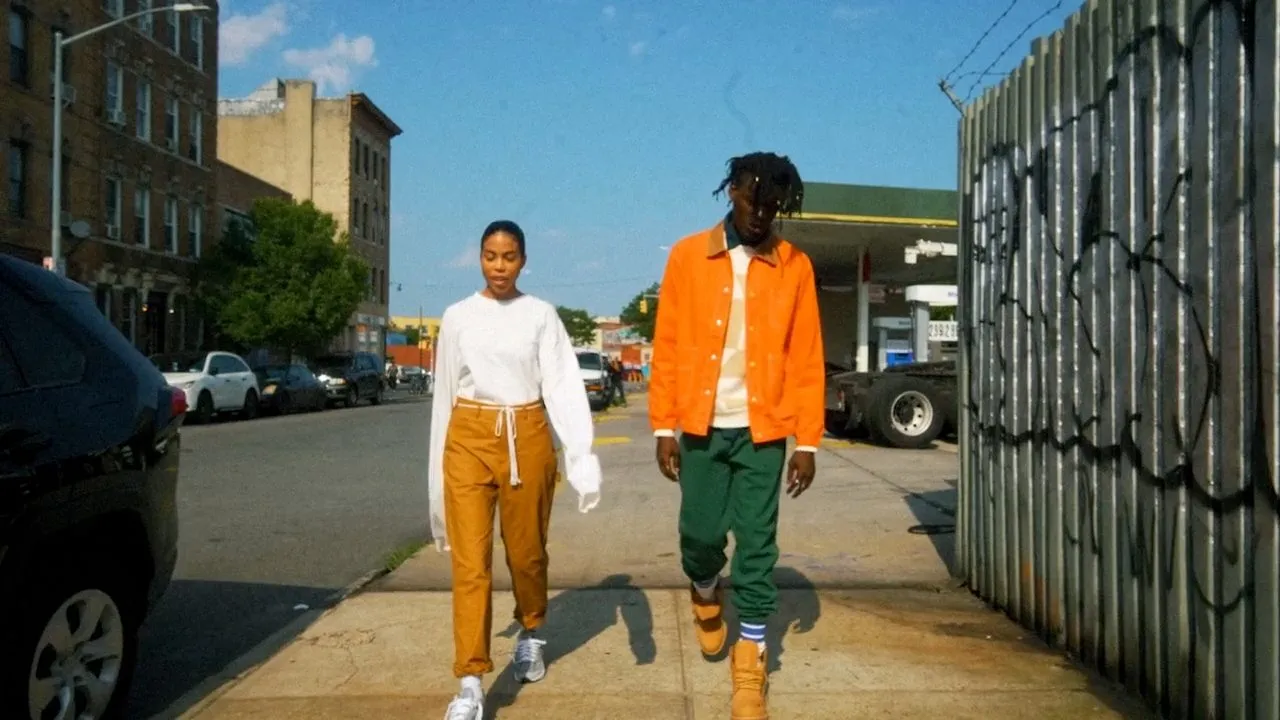 Two people walking down city sidewalk in colorful outfits, metal fencing