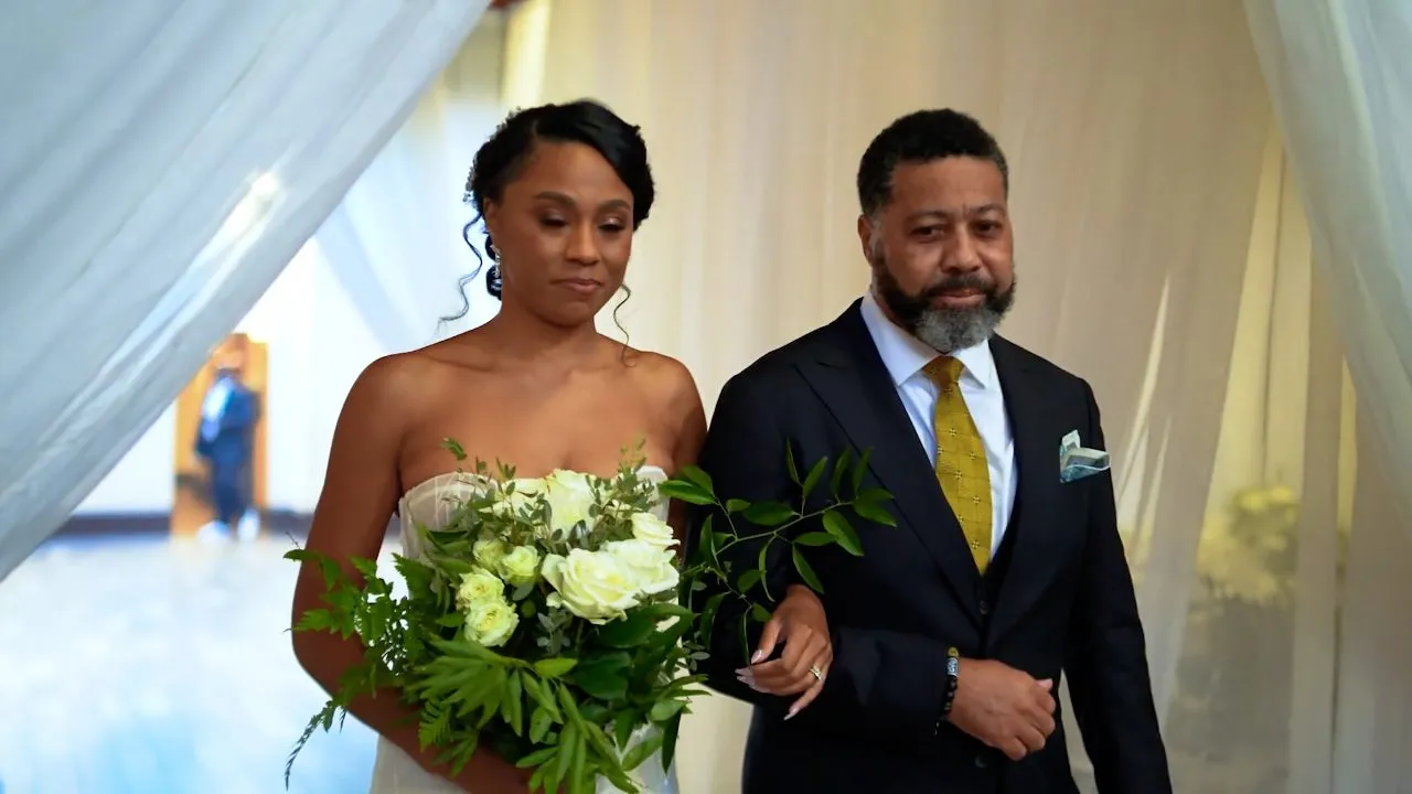 Bride holding white bouquet escorted by man in suit, draped fabric backdrop