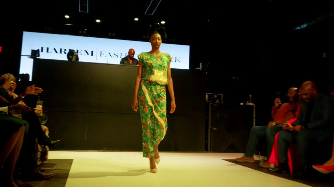 Model in green patterned dress walks runway before Harlem Fashion Week backdrop and audience