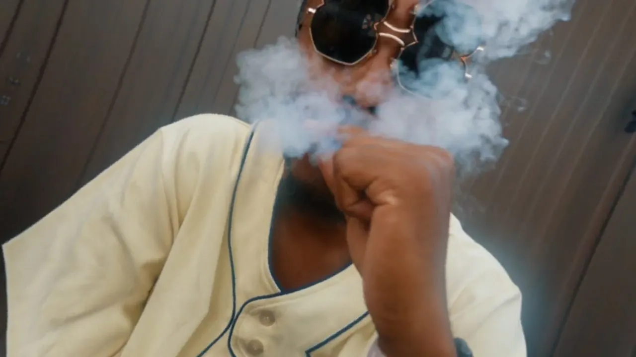 Person exhaling smoke wearing sunglasses and cream jersey, indoor dim lighting
