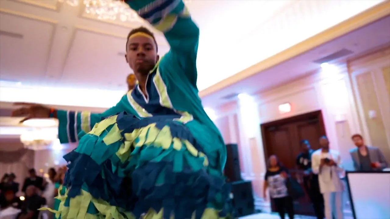 Person dancing energetically in colorful outfit at indoor reception, guests watching